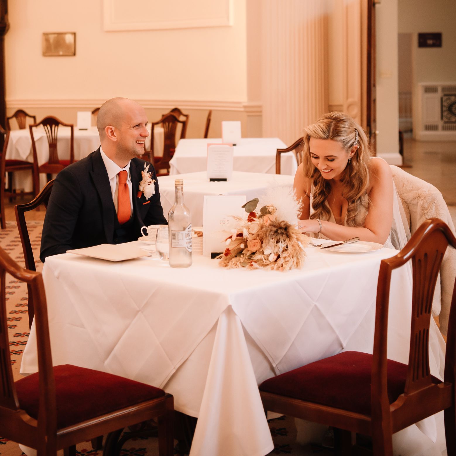 A man and a woman are seated across from each other at a small square table covered with a crisp white tablecloth. They both have happy expressions; the man is looking toward the woman with a wide smile, while she looks down at the table, laughing.