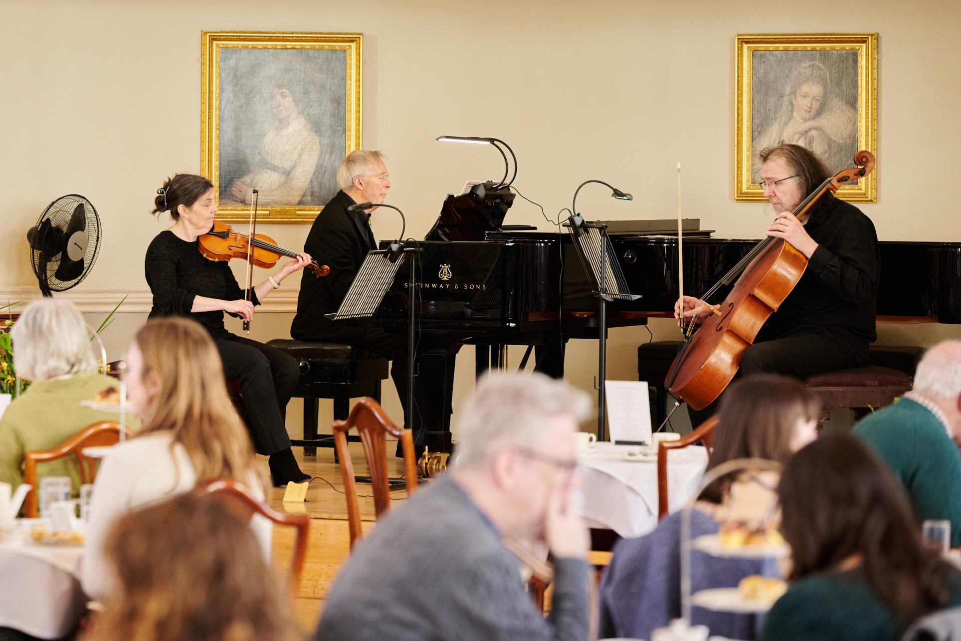 The three members of the Pump Room Trio are seated on the stage with their instruments; the cello, the violin and the piano.